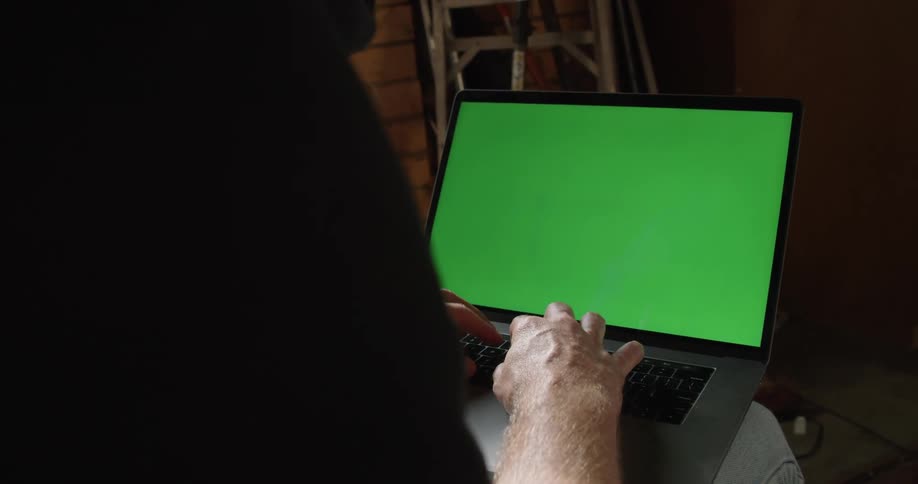 Man sitting inside a dimly lit room works on a laptop with a replaceable green screen