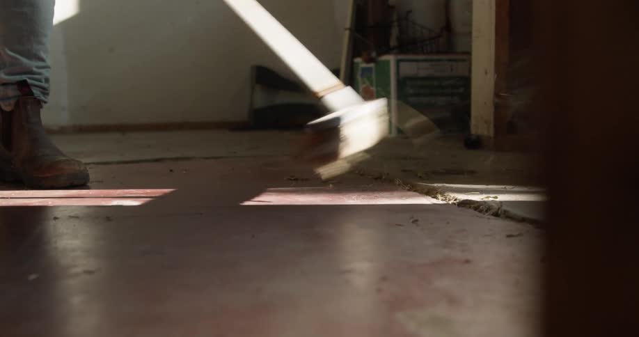 A person in a room sweeping dust with a broom with a wall and box in the background