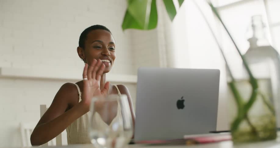 A woman sitting at a desk in front of an apple laptop waving and smiling