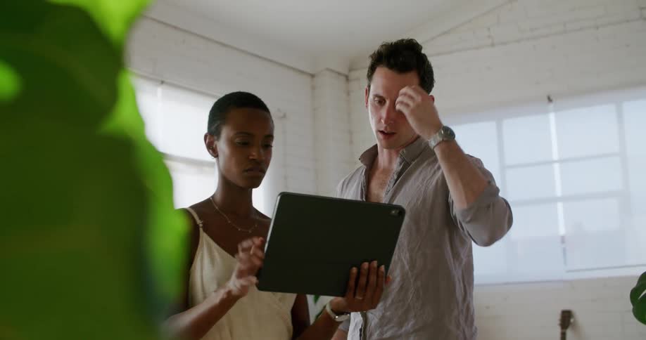 A man and a woman in a room looking at a tablet with a wall and window in the background