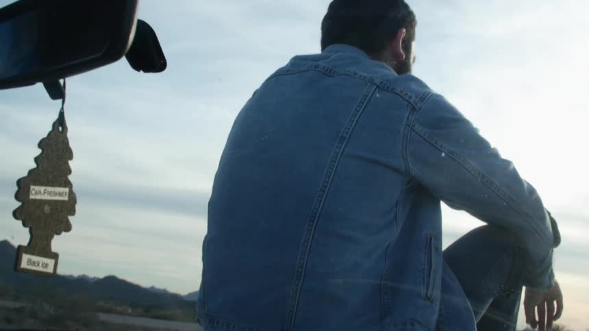 Man sits on car hood in countryside with mountains in distance