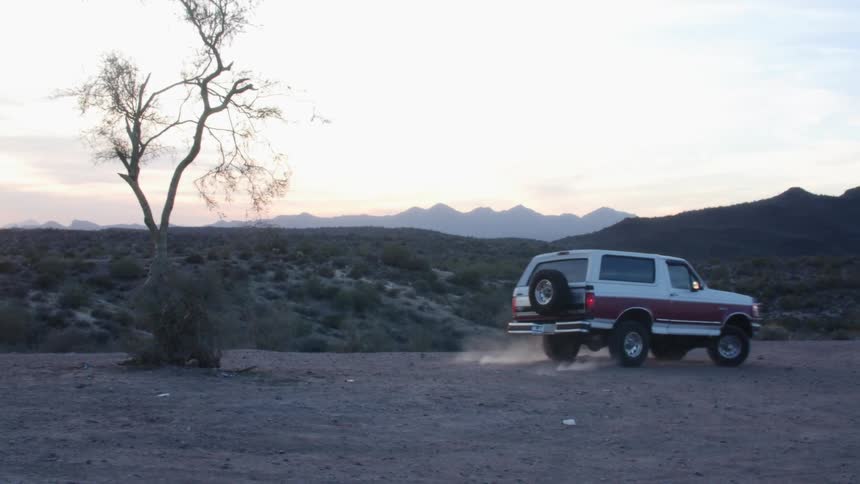 A vehicle driving in the desert with a mountain range the sky and clouds in the background