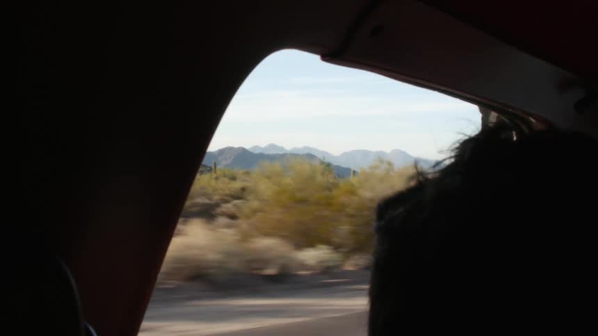 Person sits in car driving through scrub with blue mountains on horizon