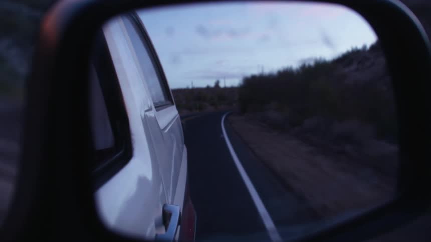 A vehicle driving with the reflection of the road the sky and clouds in the rearview mirror