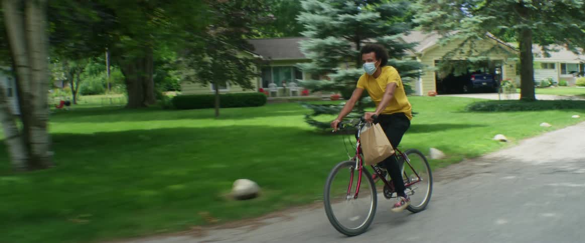 A man rides a bicycle along a road through a neighborhood while carrying a paper bag during the day