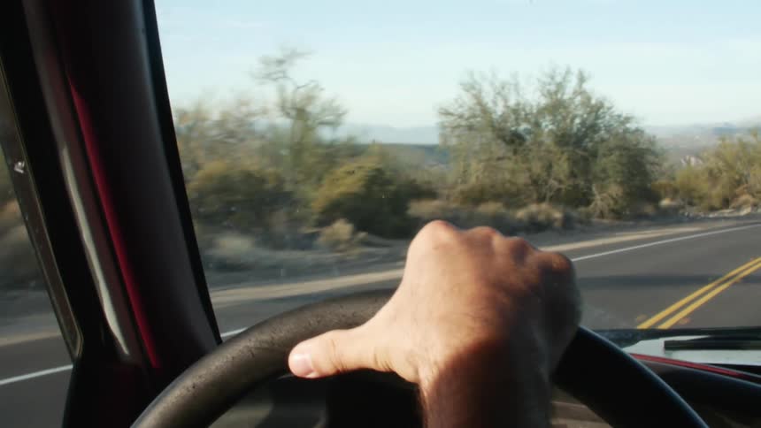A man drives a vehicle along a road through an arid countryside during the day