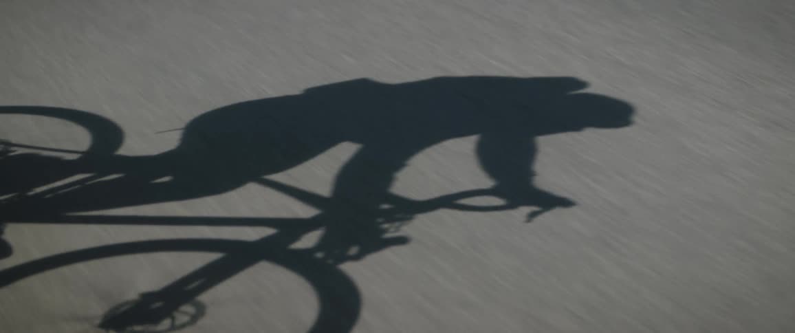A person rides their bike on a sunny day and casts a shadow onto the road that they are pedaling along