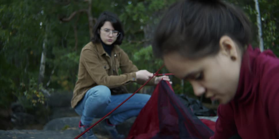 A pair of young women assemble a tent in a woodland during the day