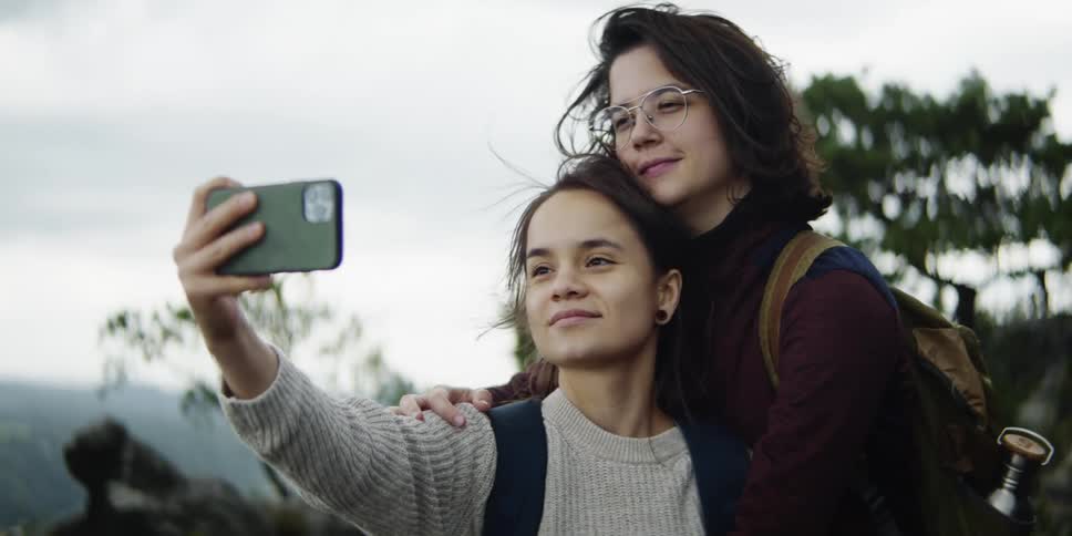 A pair of female hikers embrace for a selfie on a cloudy day
