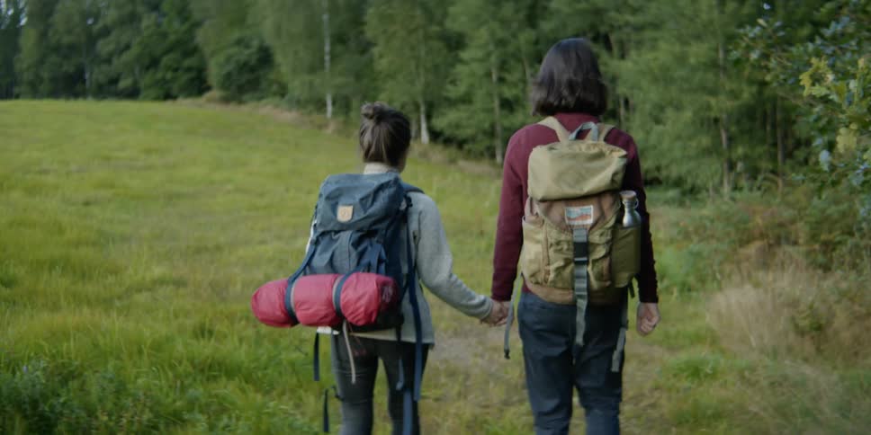 A young man and woman with backpacks walk towards a forest while holding hands