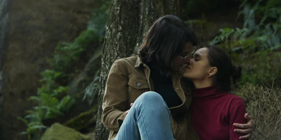 Two women sit together in a forest and share an intimate kiss