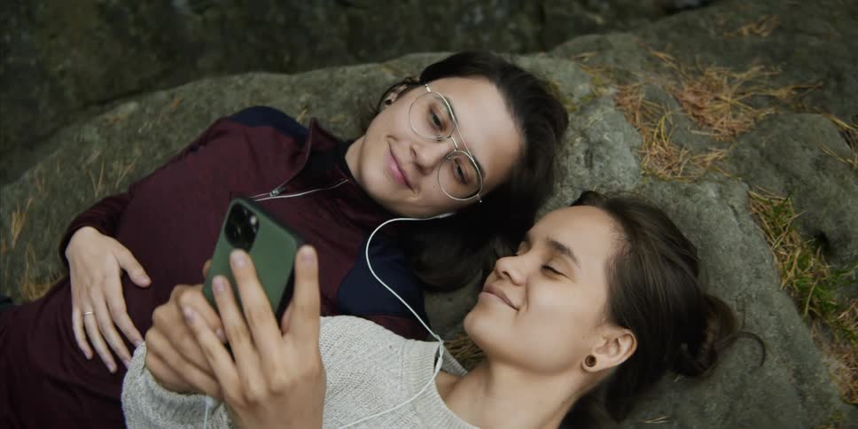 Two women lay on their backs on a rock while listening to music via shared earbuds