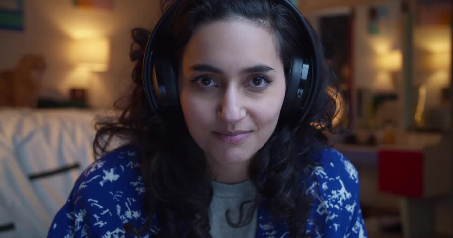 A young woman wearing headphones uses a controller to play a video game while sitting in a bedroom