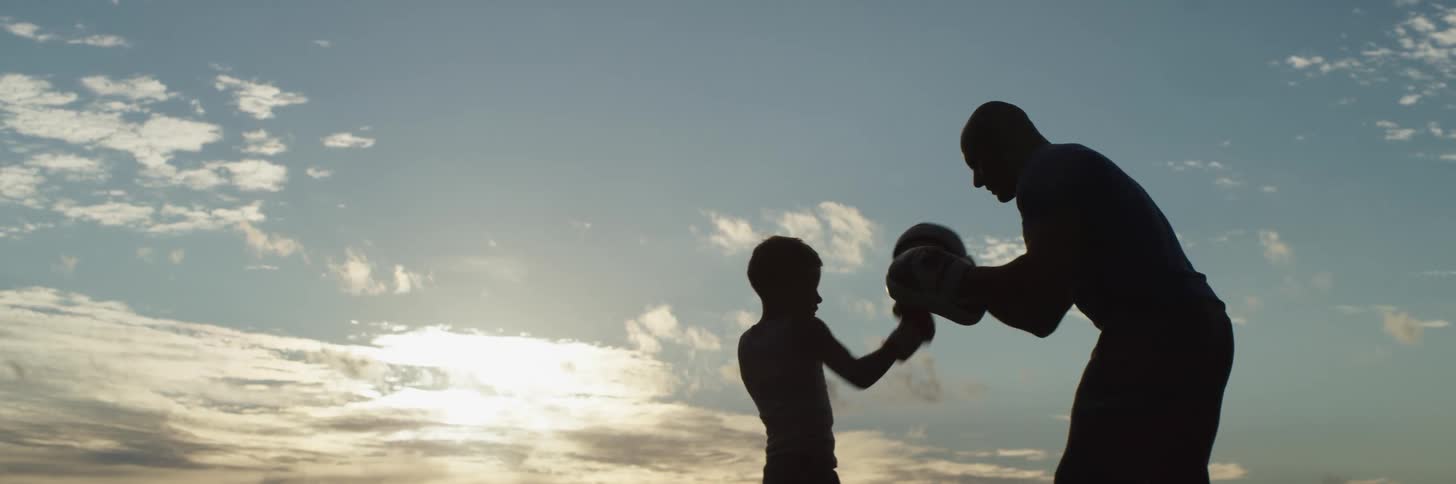 Man coaches boy in boxing standing outdoors silhouetted by the light