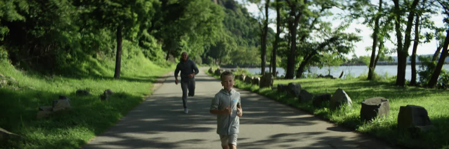 A boy and a man running on a pathway with trees a lake the sky and clouds in the background