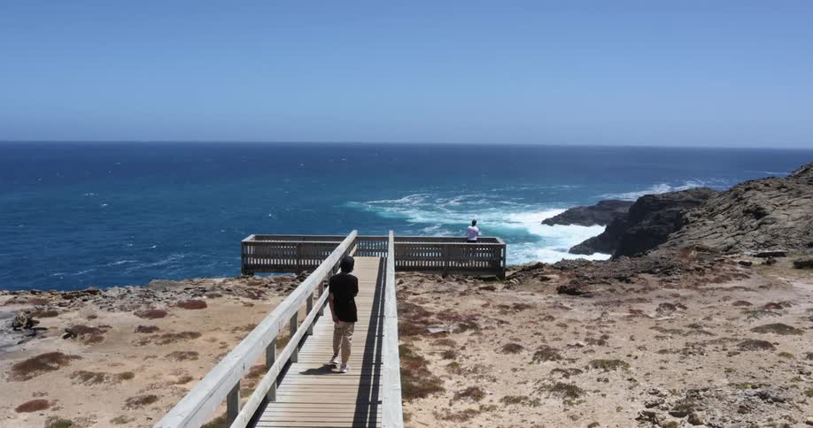 A man with a hat walking on a boardwalk with the ocean sky and clouds in the background