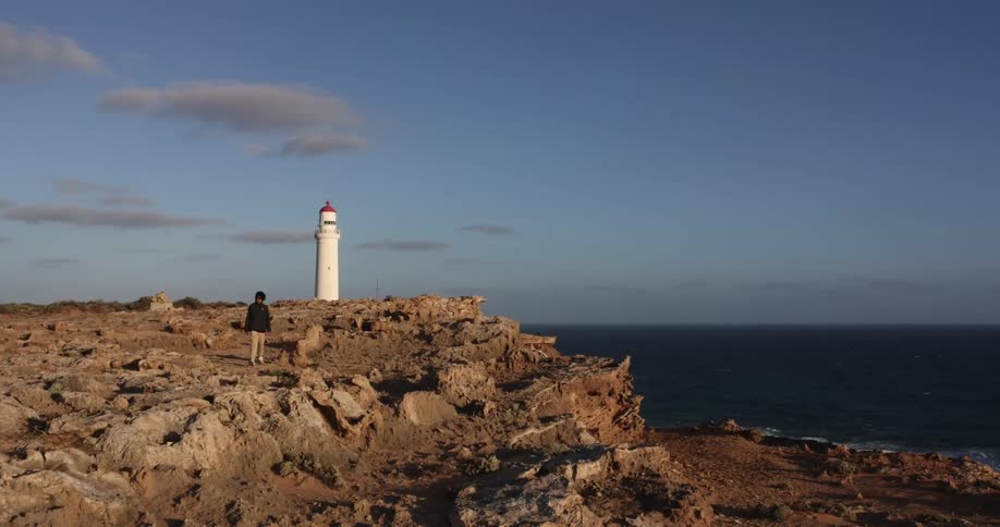 Person walking over the rocks of a cliff next to a lighthouse close to the ocean