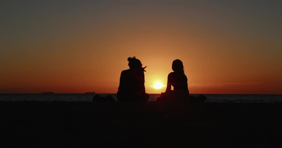 The silhouettes of two people sitting on the beach with the ocean the sunset the sky and clouds in the background