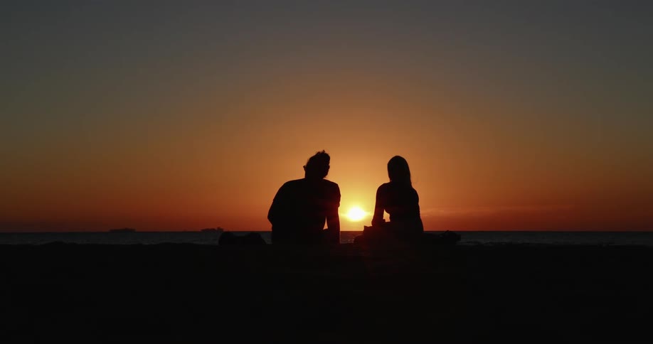 Two people sit and watch the sunset as birds fly past them