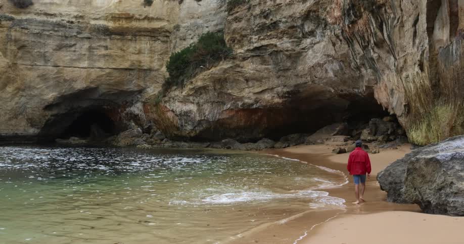 A person with a jacket walking on a beach with waves with a cliff in the background