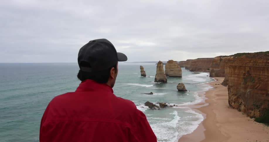 Man stands looking over beach bordered by cliff under overcast sky