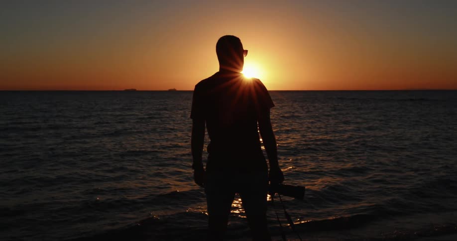 A silhouette of a man with a camera on a beach with the ocean the sunset the sky and clouds in the background