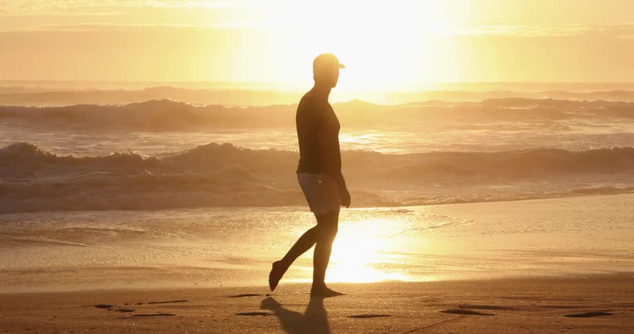 A man in a baseball cap walks along the beach as the sun tints the ocean and sky a pale yellow