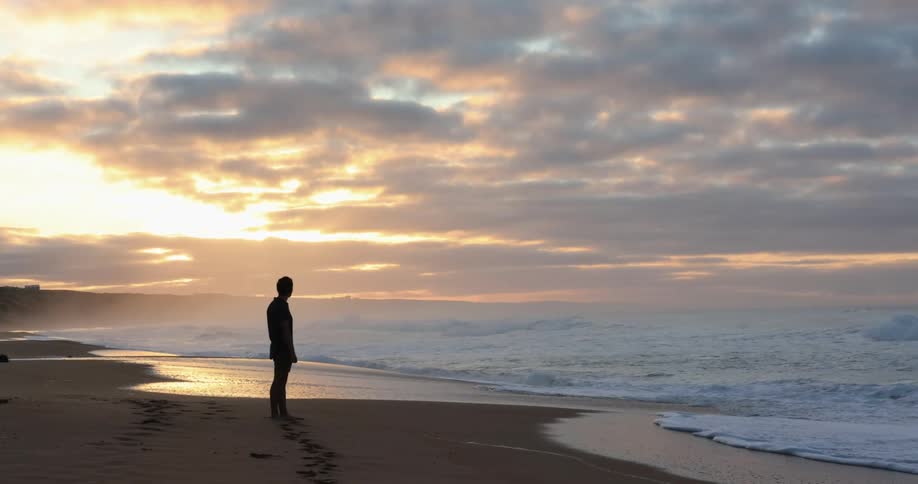 A man stands alone on an empty beach as waves crash onto the shore at sunset