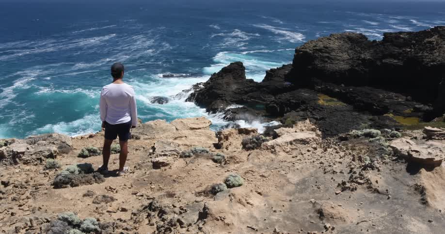 A person with a hat standing on a rocky coast with waves
