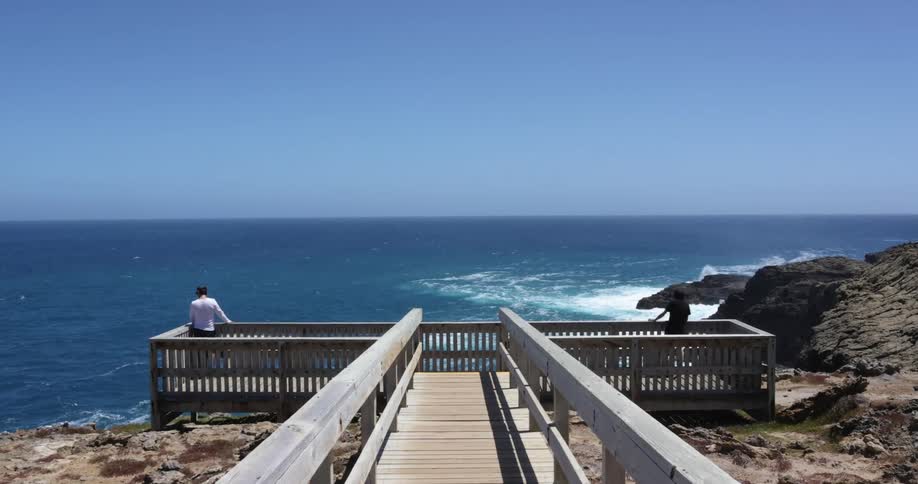 Woman and man standing on the opposite sides of a wooden balcony looking over the ocean