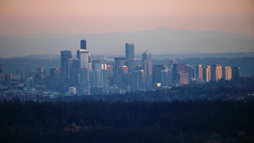 Sunset light falls on the skyscrapers of downtown Seattle as the hills of the Cascade mountains rise in the background in this aerial