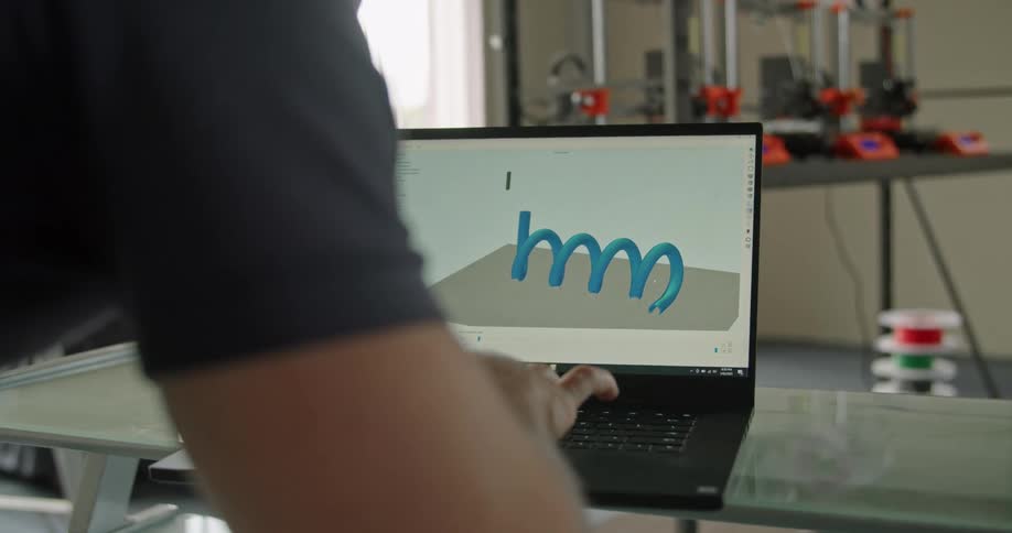 A person using a laptop with a 3d printer a wall and window in the background