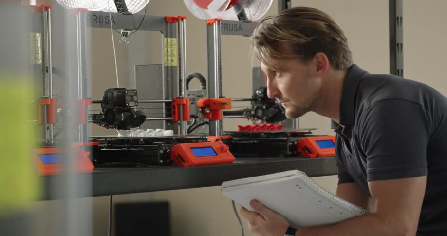 A man takes notes in a notebook while observing a table of 3d printers