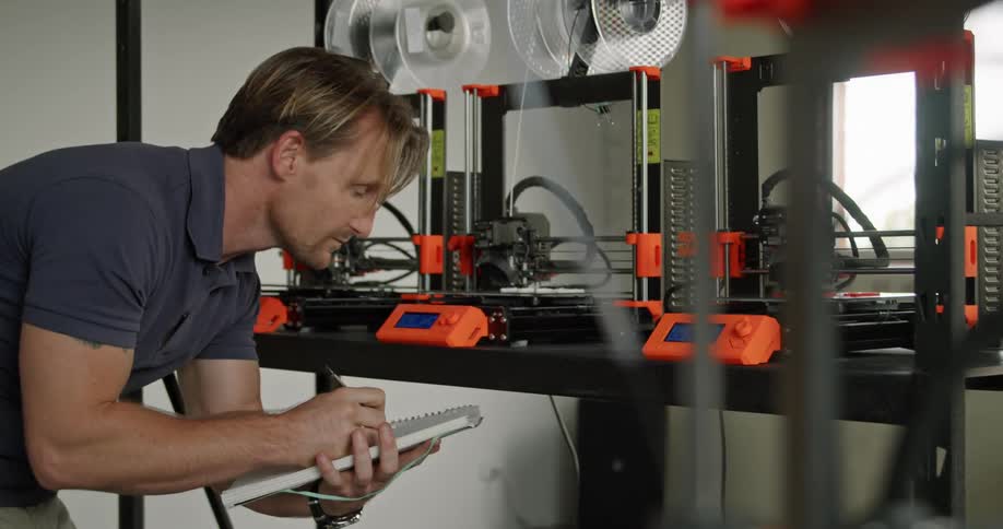 A man with a pen and a notebook looking at a 3d printer with a wall and window in the background