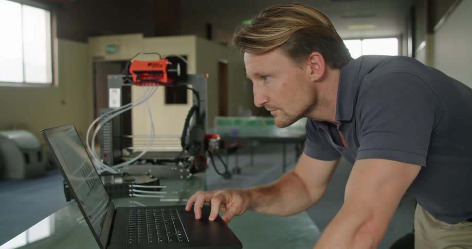 A man leans over a glass table while looking at a graph on a laptop attached to electrical equipment
