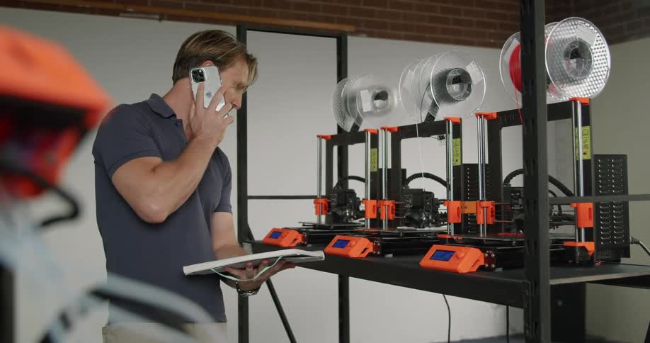 A man holding a notebook answers his phone and talks as 3d printers print