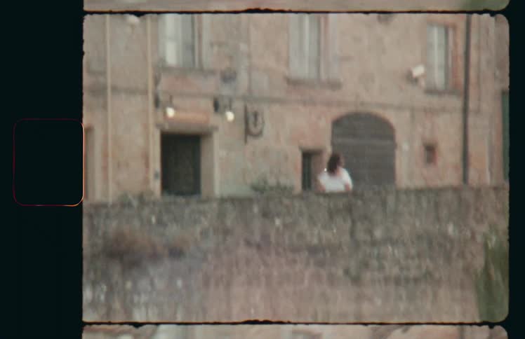 Woman waves at camera operator before walking along between wall and apartment building