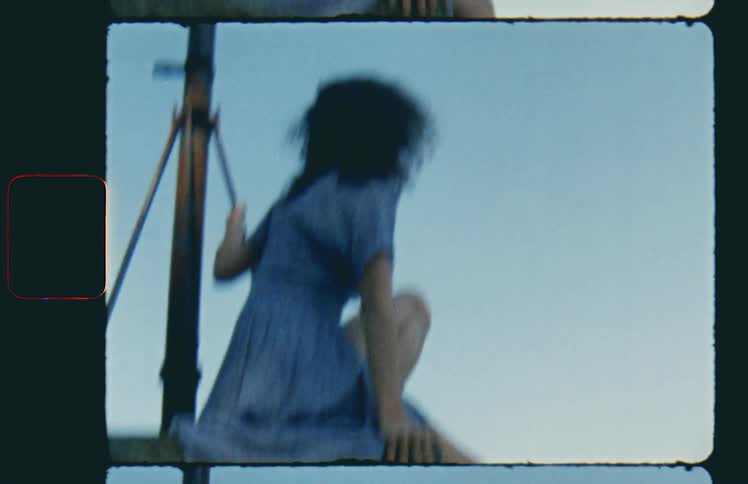 A white woman in a blue dress sits high up on stones and holds on to a metal pole while looking into the distance