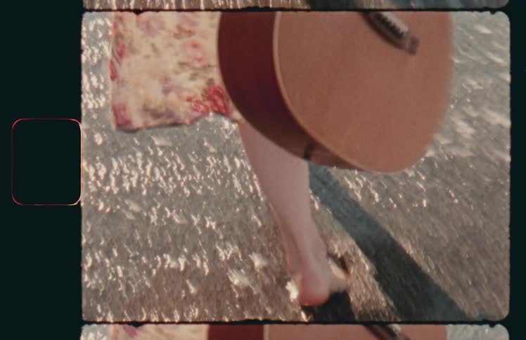 A woman carrying a guitar walks through the water on a beach