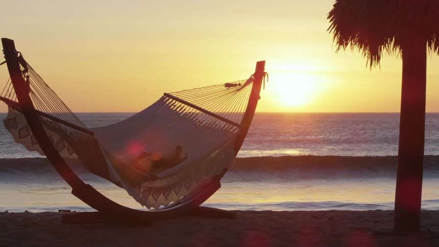 A person rests on a hammock on a beach at sunset