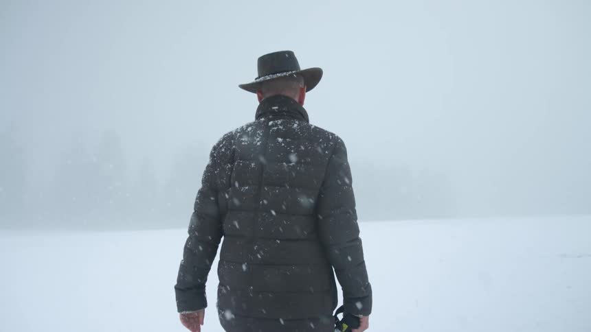 Snow falls as a man walks through a snow covered field while holding a large camera