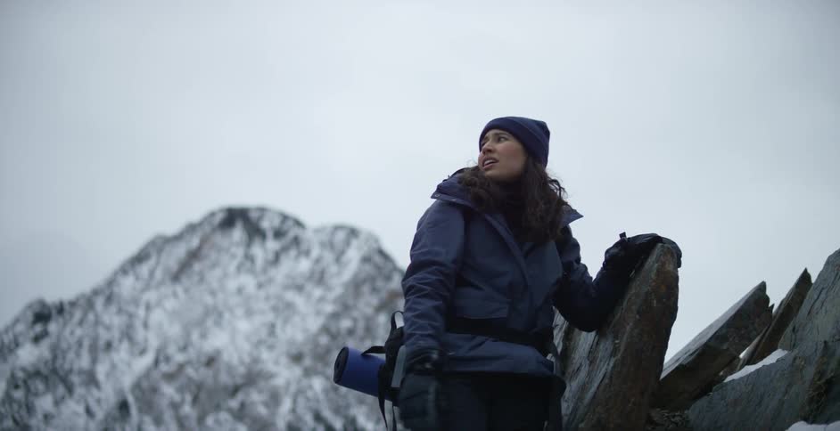 A woman climbing onto a rock while hiking in snowy mountains on an overcast day