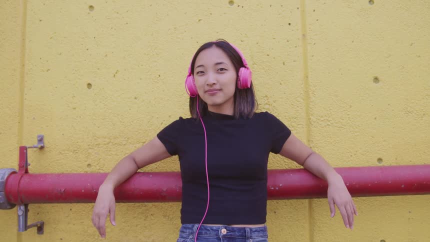 Young girl wearing pink headphones reclined on a red pipe against a yellow wall looking at the camera