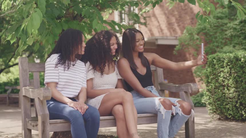 Three female friends smiling and posing for a selfie on an outdoor park bench