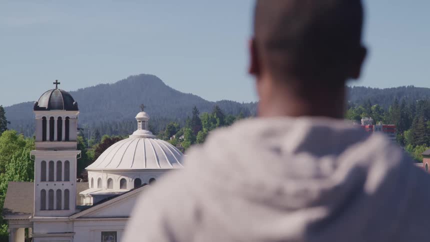 A man with a hoodie standing with a church trees the sky and clouds in the background