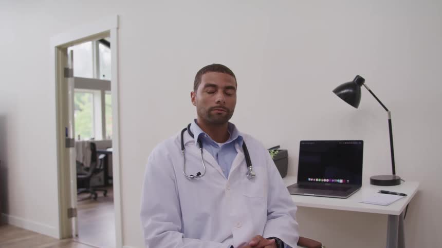 A doctor sitting with his back to a laptop turns his head to look into the camera
