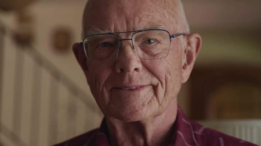 An elderly man with eyeglasses blinking and smiling indoors