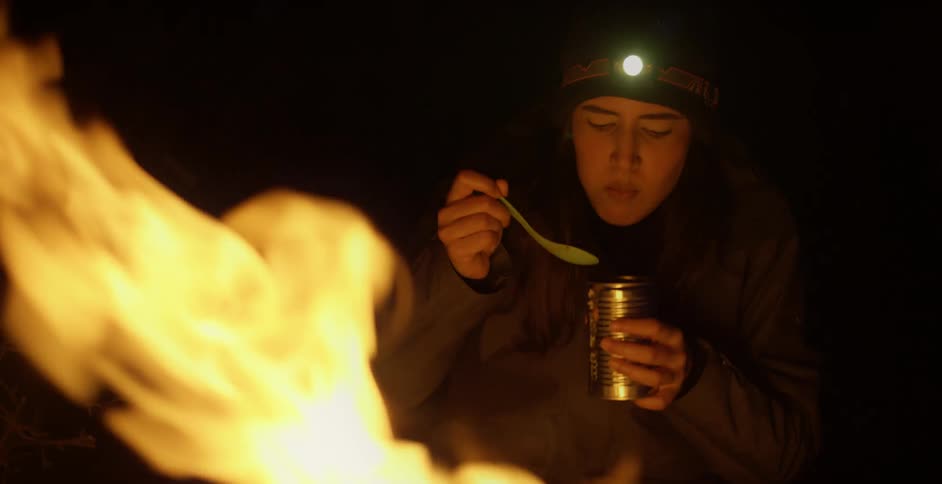 A teenage boy sitting by a fire eats out of a can