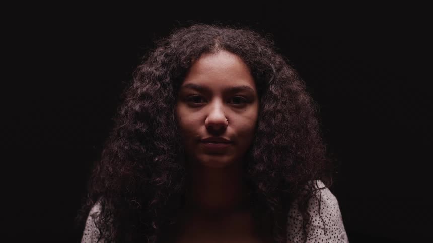 A woman lifts her head and looks straight ahead against a black background