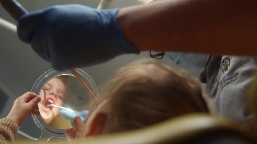 A young girl lies in a dental chair and holds up a mirror during a procedure